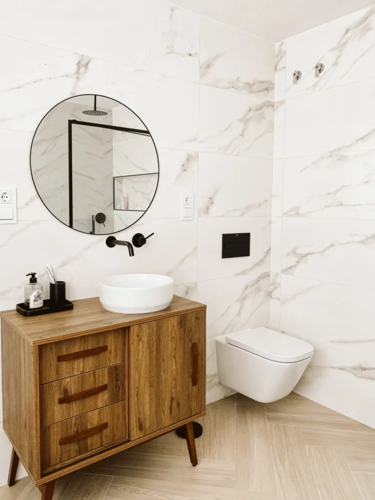 Lavabo sobre mueble de madera, espejo circular y grifería negra sobre un revestimiento blanco veteado.